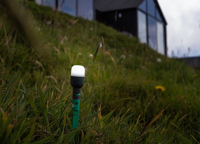 A green garden light stands in tall grass, with a modern black building in the background under a cloudy sky.