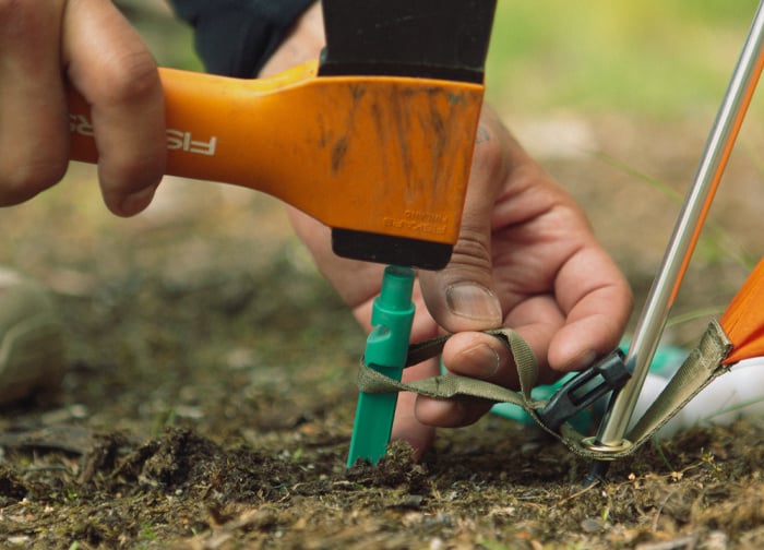 A hand is using an orange hammer to drive a green tent stake into the ground, securing a tent.