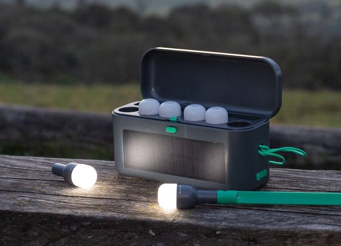 A portable speaker with a solar panel, open lid, and two small lanterns on a wooden surface outdoors.