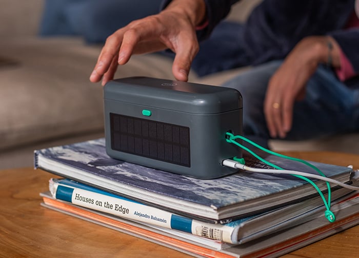 A person is reaching for a portable speaker on top of stacked books, with a charging cable connected.