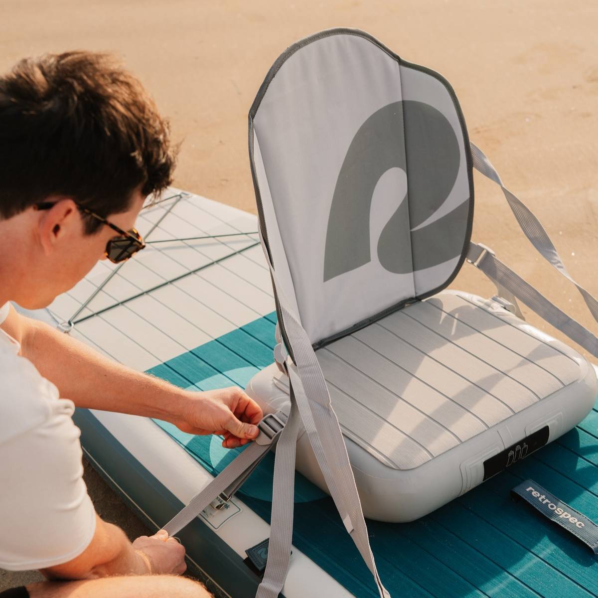 A person adjusts the straps of a comfortable inflatable kayak seat attached to a paddleboard on a sandy beach under warm sunlight.