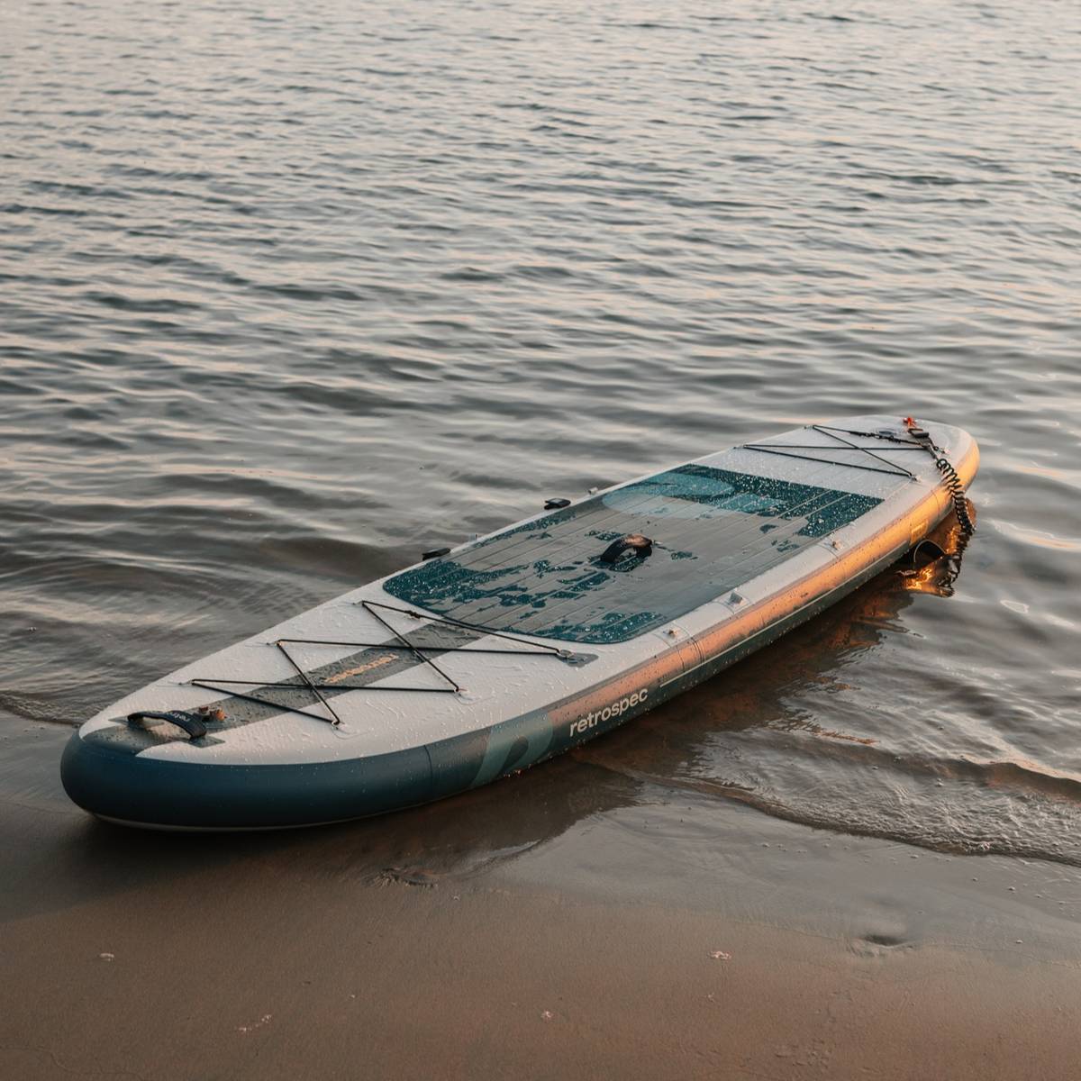 An Weekender Plus Inflatable Stand Up Paddle Board Kayak Hybrid 10'6" rests on the shore, partially in calm water, reflecting soft evening light. Gentle waves lap at the sand.