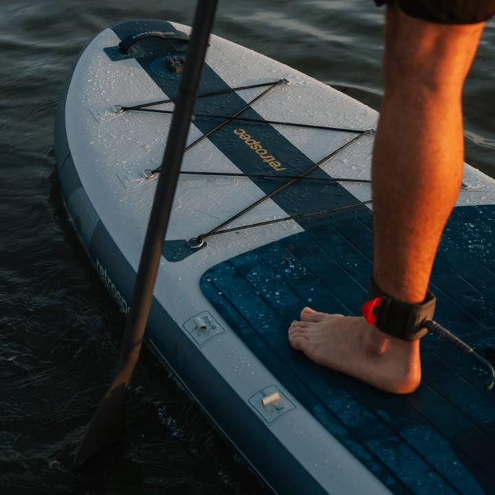 A person's foot on a paddleboard, with a paddle in the water and droplets glistening on the board in the evening light.