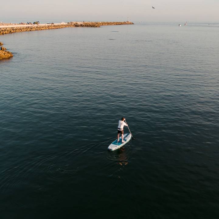 A person paddleboards on calm waters, with a rocky jetty and beachgoers visible in the distance under a clear sky.