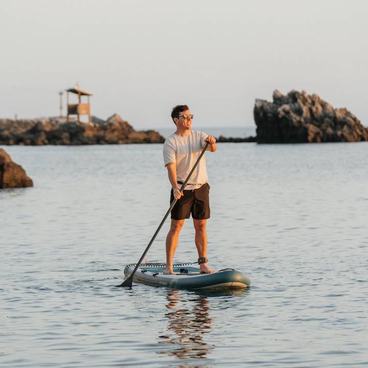 A person paddleboarding on calm water, with rocky formations and a small structure in the background during sunset.
