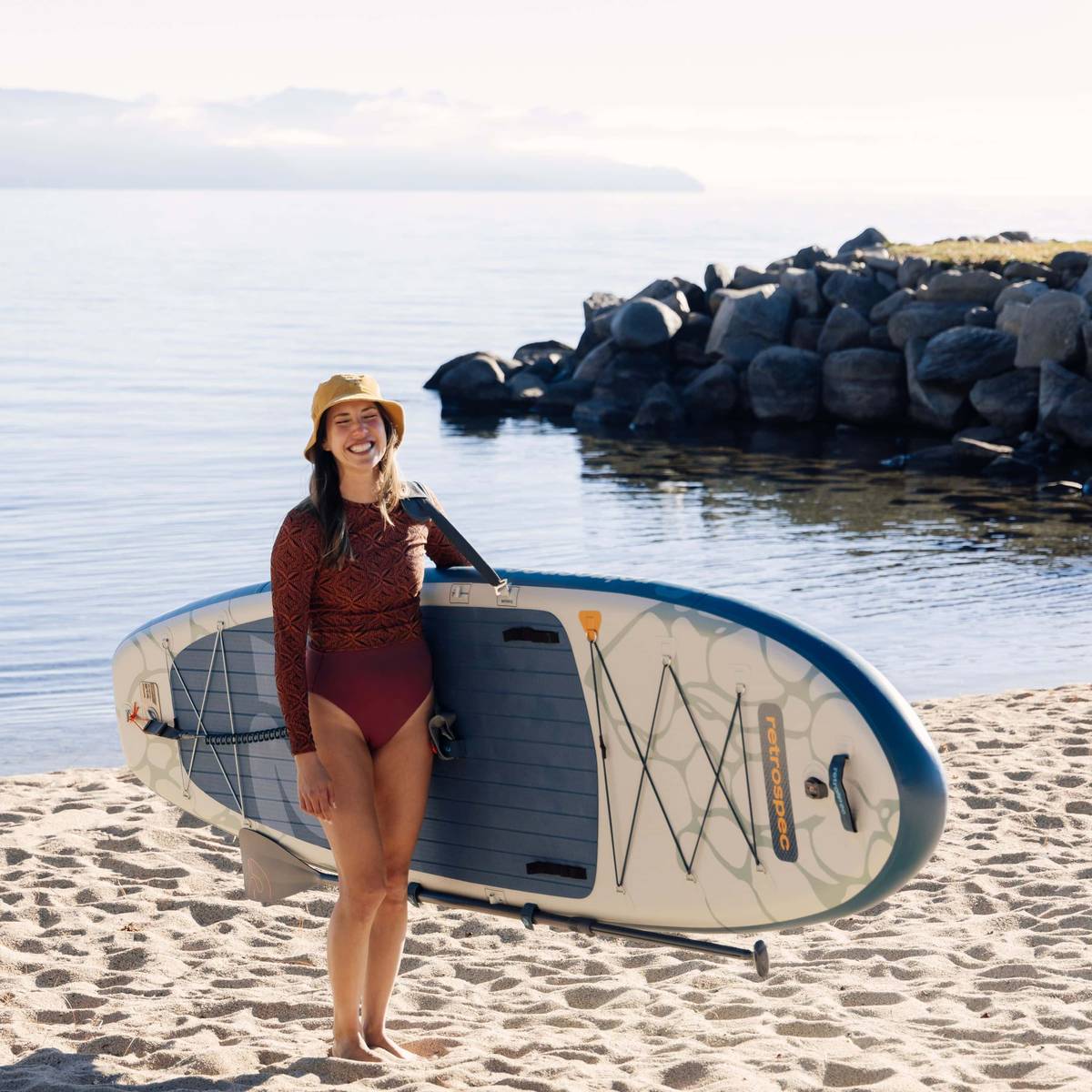 A person stands on the beach holding an inflatable paddleboard, wearing a red long-sleeve top and high-waisted bikini, with rocks and water in the background.