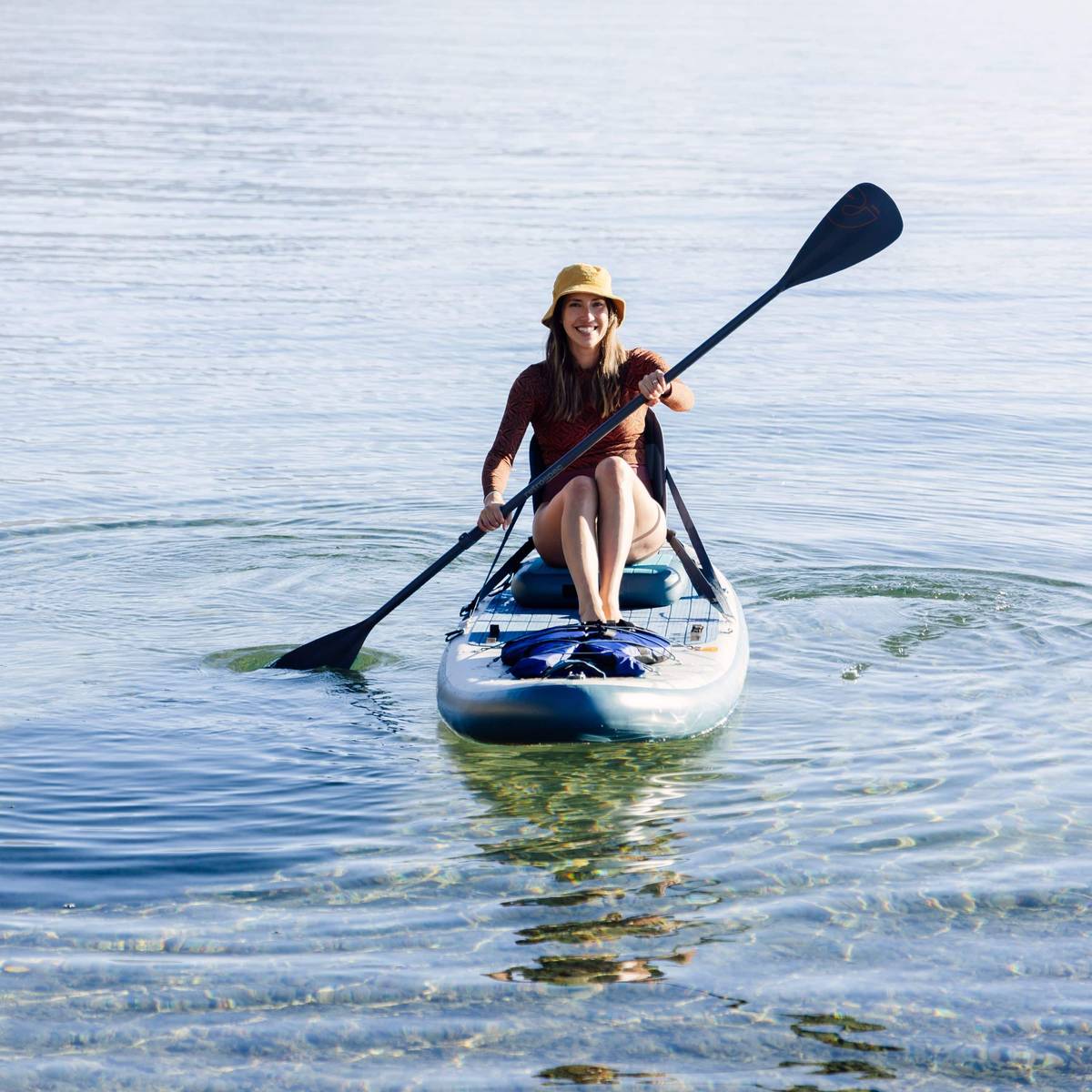 A person paddles an inflatable stand up paddle kayak hybrid on calm, clear water, wearing a sun hat and enjoying a serene outdoor experience.
