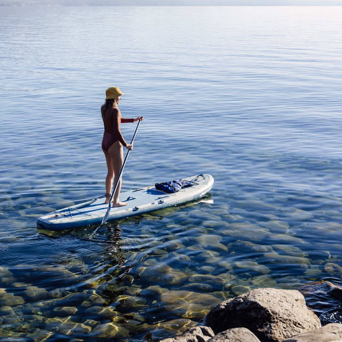 A person paddles an inflatable stand up paddle on calm, clear water, wearing a sun hat and enjoying a serene outdoor experience.