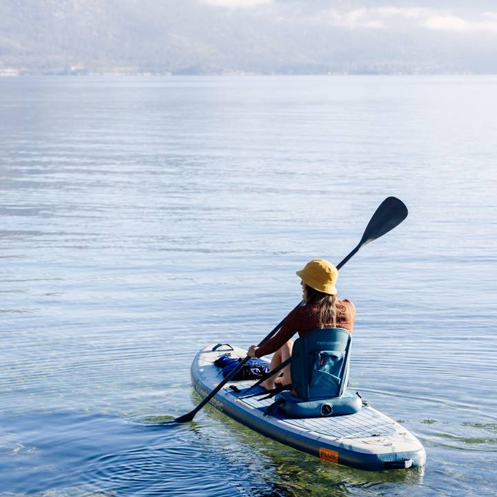 A person paddles an inflatable stand up paddle kayak hybrid on calm, clear water, wearing a sun hat and enjoying a serene outdoor experience.