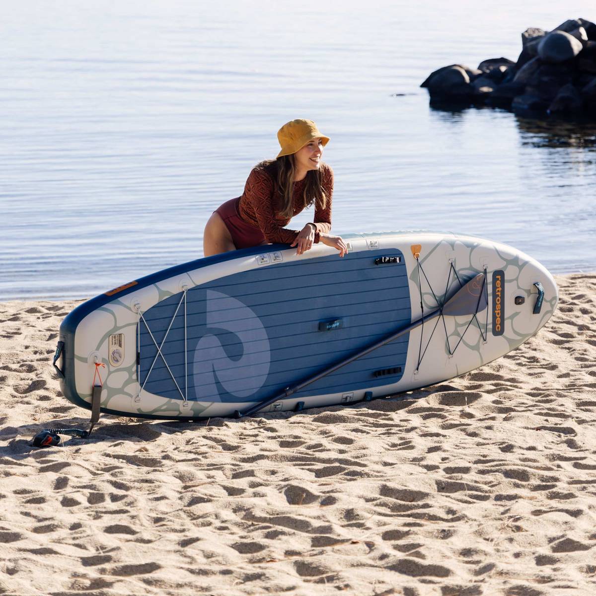Person in a swimsuit kneeling by an inflatable paddleboard on a sandy beach, with calm water and rocks in the background.