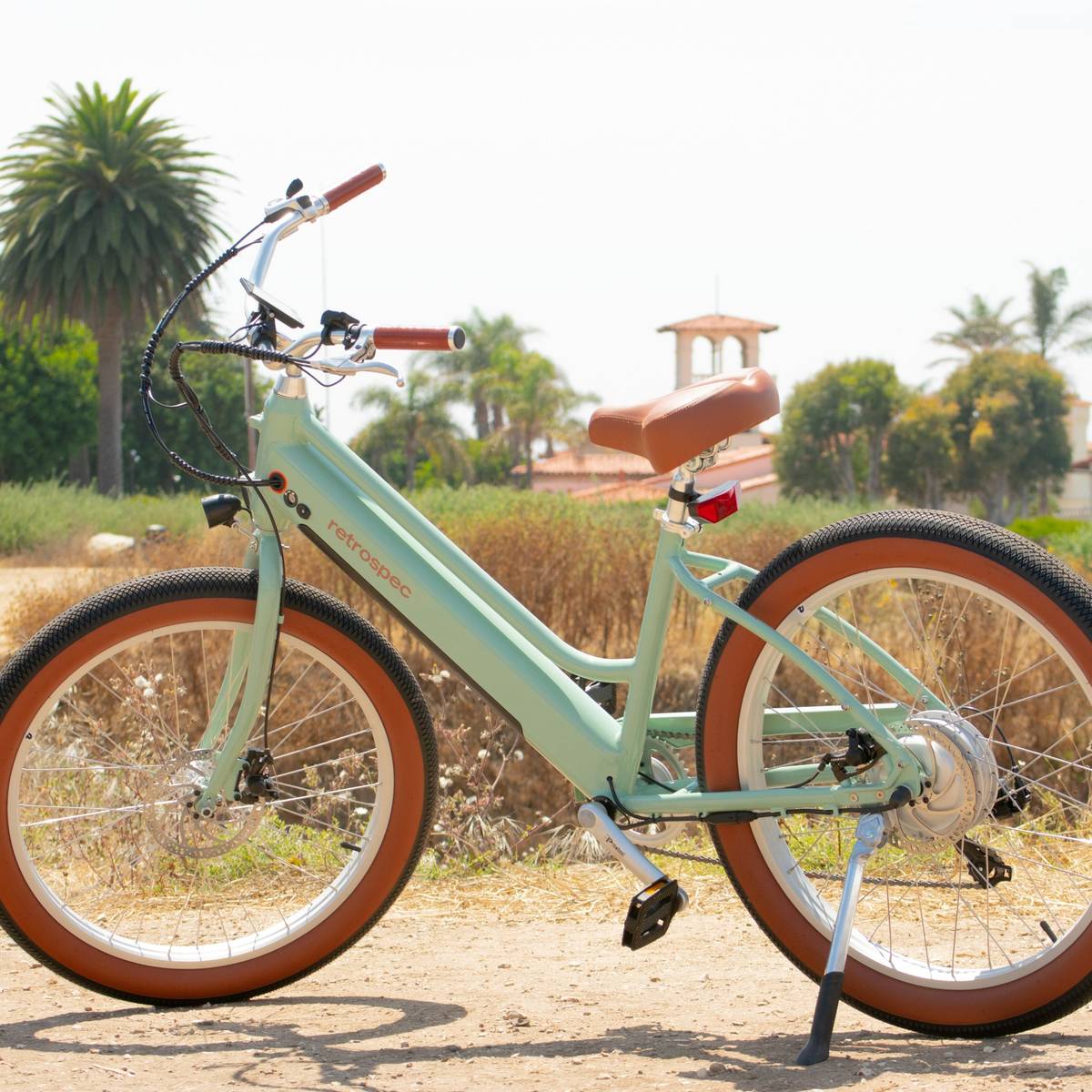 A mint green Chatham Rev+ Electric Beach Cruiser Bicycle with brown accents stands on a dirt path, surrounded by greenery and a sunny, bright sky in the background.