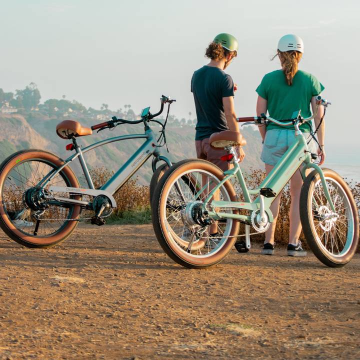 Two electric bicycles parked on a sandy path overlooking a scenic coastline, with lush cliffs and palm trees in the background.