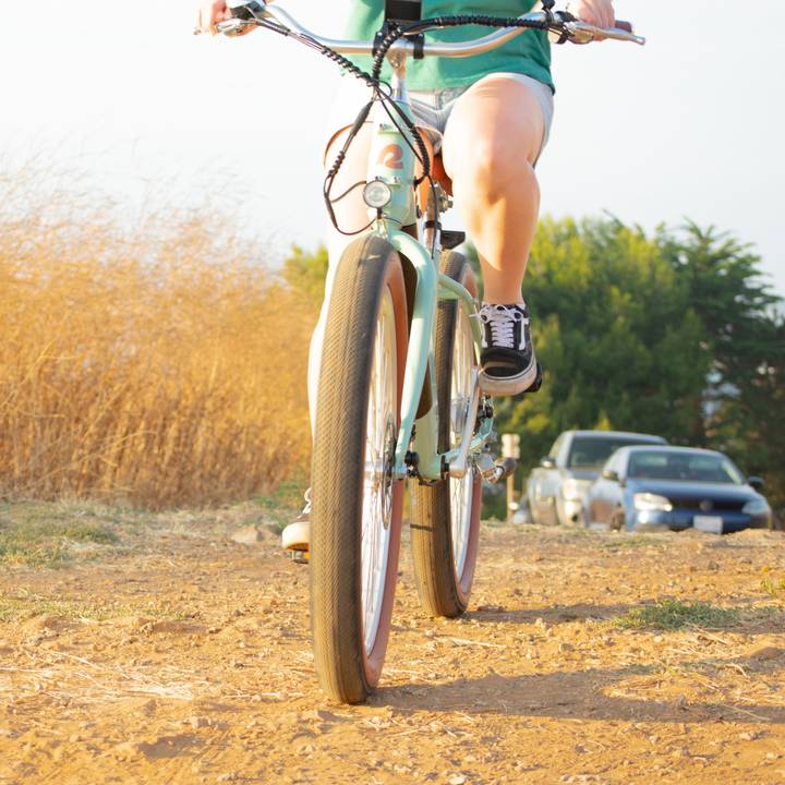 A person rides a mint-green Chatham Rev Plus Electric Beach Cruiser Bike on a dirt path, surrounded by tall grass and parked cars in the background during golden hour.