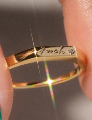 A close up of a gold ring engraved with the word fuck pinched between two fingers