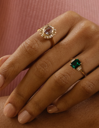 A close up of two gold gemstone rings on a hand in front of a pink background