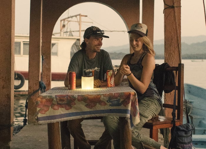 A man and a woman sit at a table on a dock, enjoying drinks and a warm conversation, with a scenic view in the background.