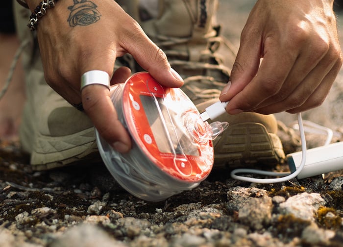 A person is connecting a charging cable to a round, red device on the ground, near their beige shoes.