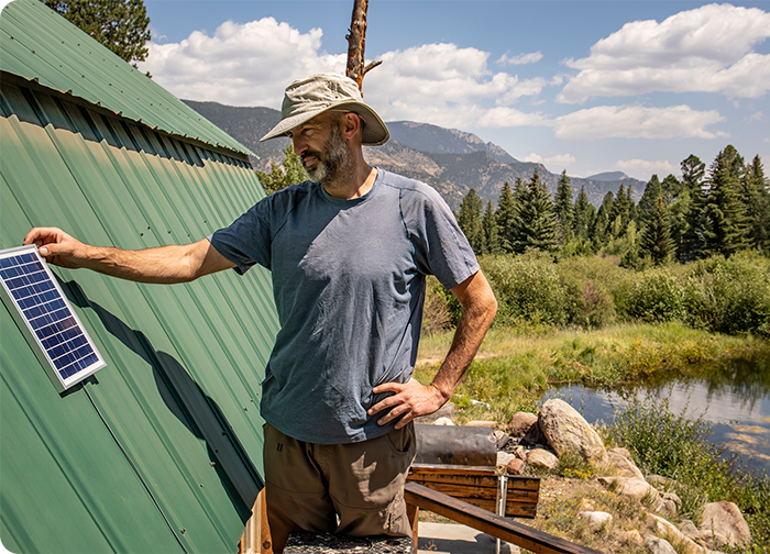 A man adjusts a small solar panel on a green metal roof, surrounded by trees and mountains under a blue sky.