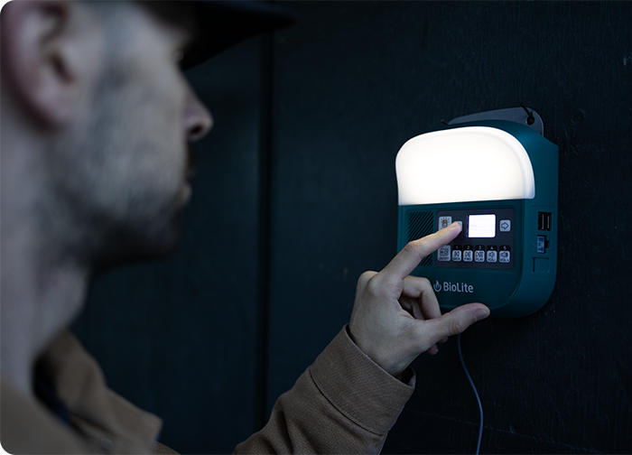 A person interacts with a BioLite device mounted on a wall, adjusting settings on its illuminated control panel.