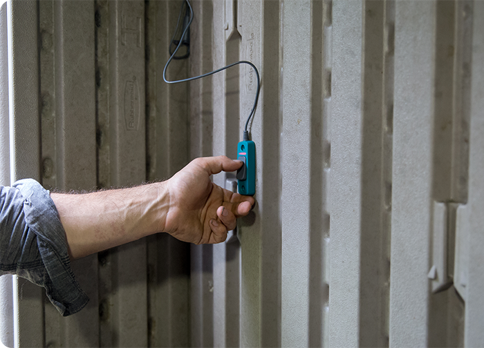 A hand is pressing a blue device against a textured wall, connected by a wire.