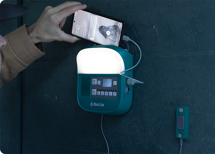 A person connects a smartphone to a teal BioLite device mounted on a wall, featuring a bright light and control panel.