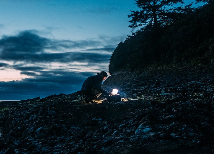 A person sits on a rocky shore at dusk, illuminated by a laptop, with trees and clouds in the background.