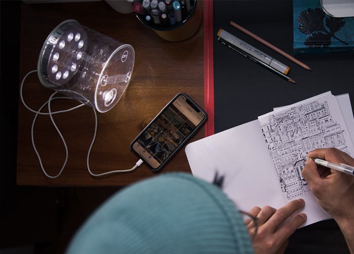 A person wearing a beanie sketches on paper, with a phone displaying images and a lamp illuminating the workspace.