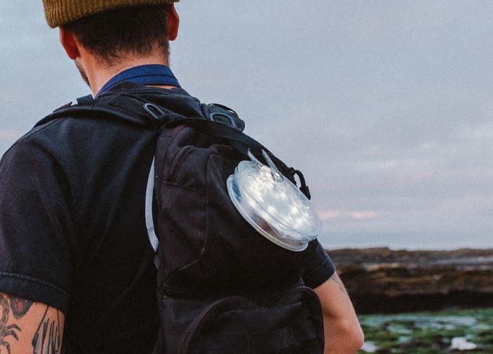 A person with a backpack stands outdoors, wearing a beanie, with a circular light attached to the side of the pack.