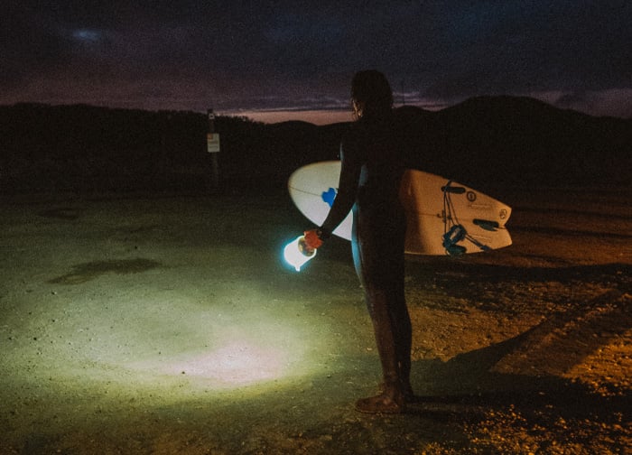 A surfer in a wetsuit holds a lantern, standing on a dark beach with a surfboard beside them, illuminated by the light.