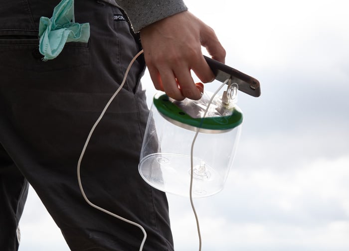 A person holds a transparent container with a green lid and a cable, standing outdoors against a cloudy sky.