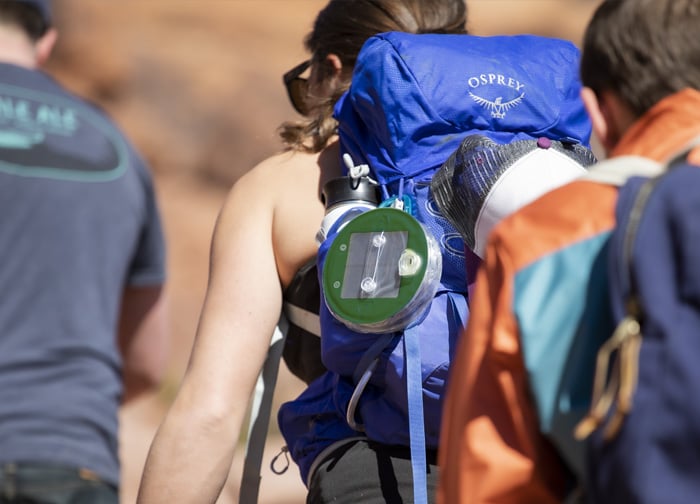 A person wearing a blue Osprey backpack with a hydration system, walking with others in a natural outdoor setting.