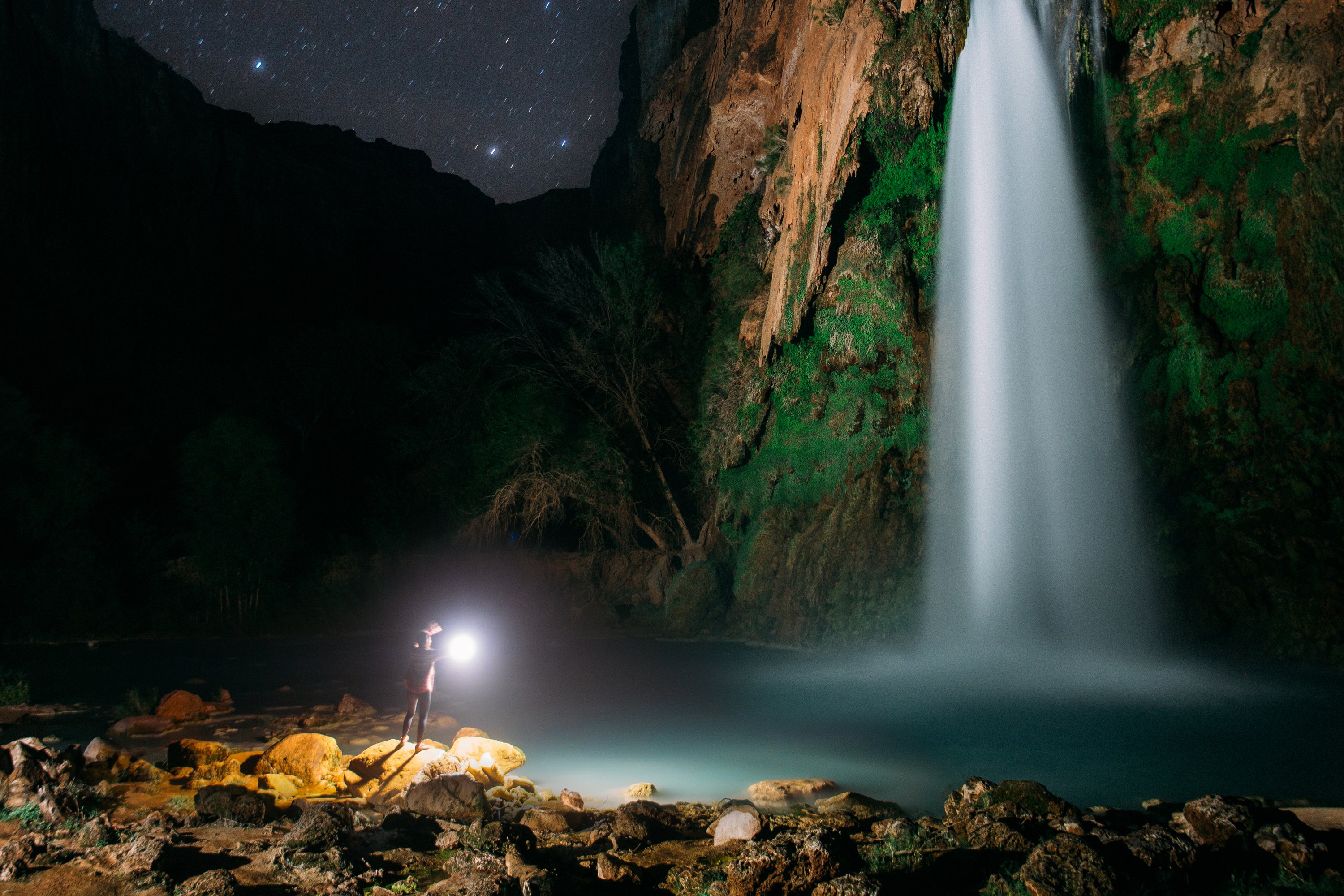 A person stands on rocks by a waterfall at night, holding a light. Stars twinkle in the dark sky above.