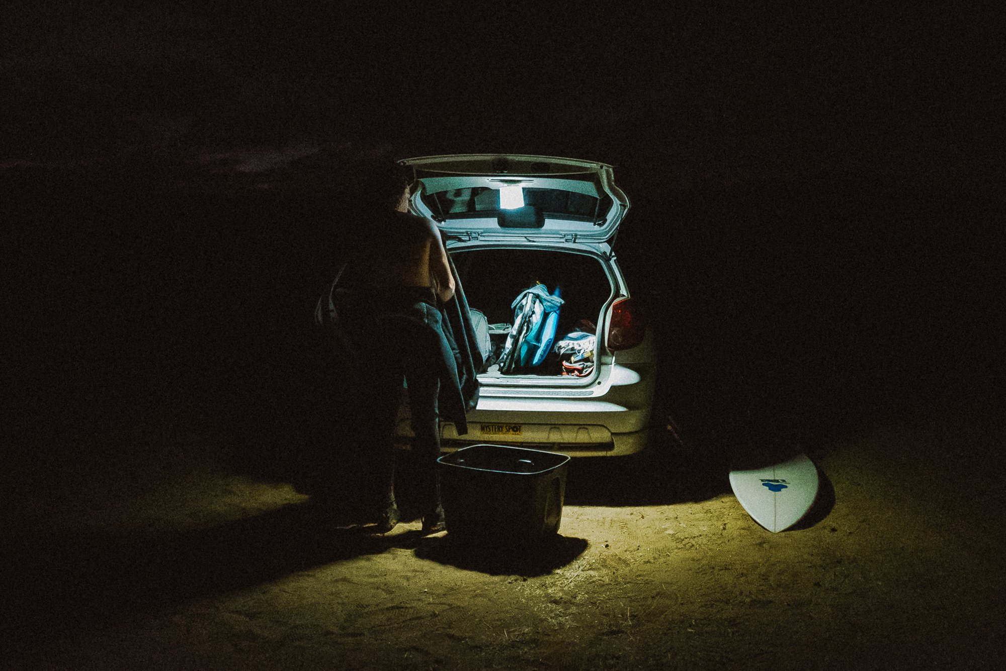 A person stands by the open trunk of a car at night, illuminated by a light, with a surfboard and bags nearby.
