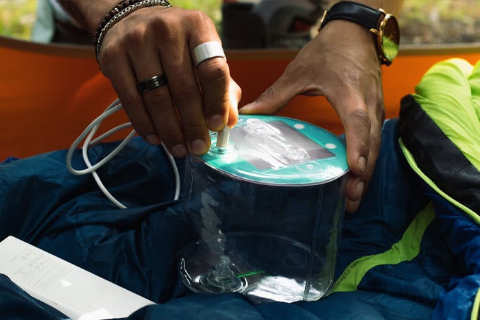 A person is using a device with a clear container and a hose, possibly for inflating or pumping, on a blue sleeping bag.