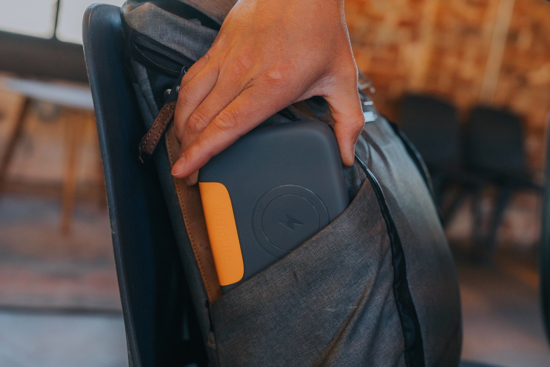 A hand is placing a portable charger into the pocket of a backpack, set against a rustic background.