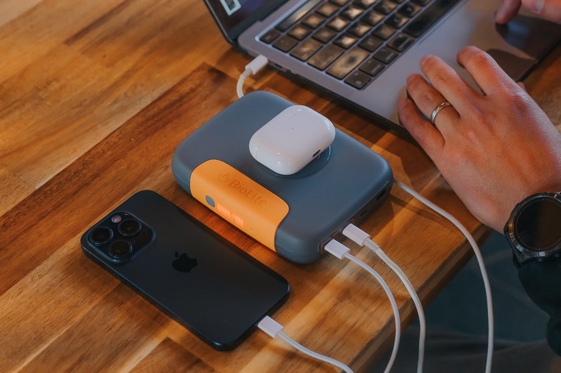 A smartphone, AirPods case, and a power bank with charging cables are on a wooden table, next to a laptop.