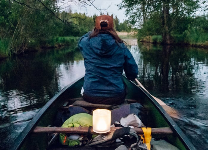 A person paddles a canoe on a calm waterway, surrounded by trees, with a lantern glowing on the boat.