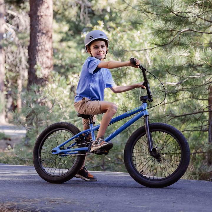 A child in a blue shirt and helmet rides a blue Sesh 20" Youth BMX Bike on a paved path surrounded by greenery.