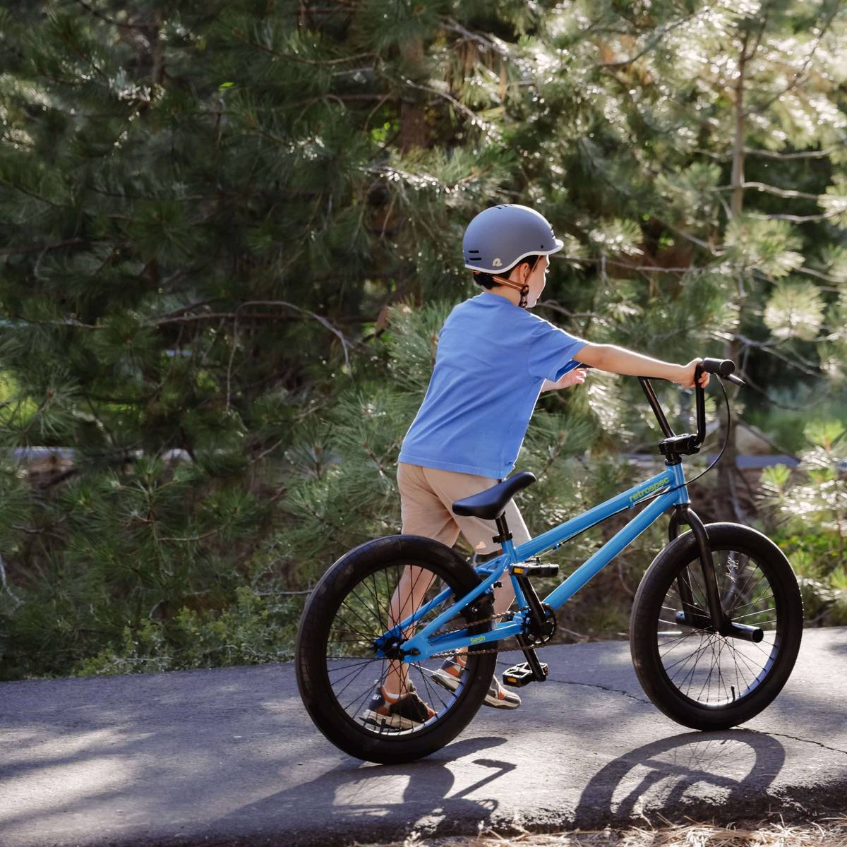 A young boy in a blue shirt and helmet walks beside his blue Sesh 20" Youth BMX Bike on a path, surrounded by lush green trees.