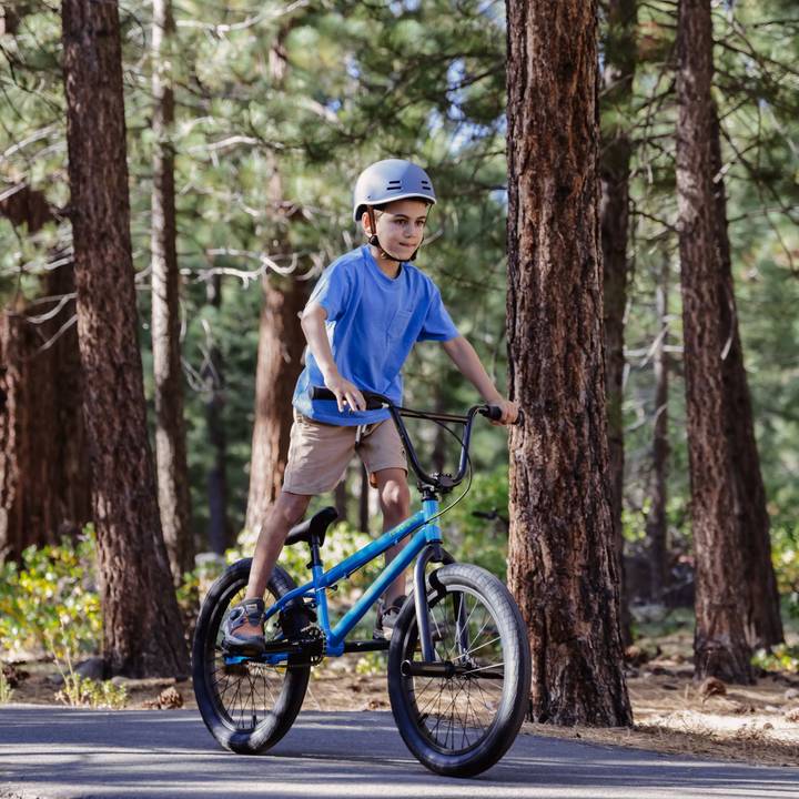 A child rides a blue Sesh 20" Youth BMX Bike on a forest trail, surrounded by tall trees and greenery, wearing a helmet for safety.