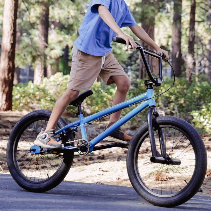 A child rides a blue Sesh 20" Youth BMX Bike on a forest path, wearing a blue shirt and khaki shorts, with trees and greenery in the background.