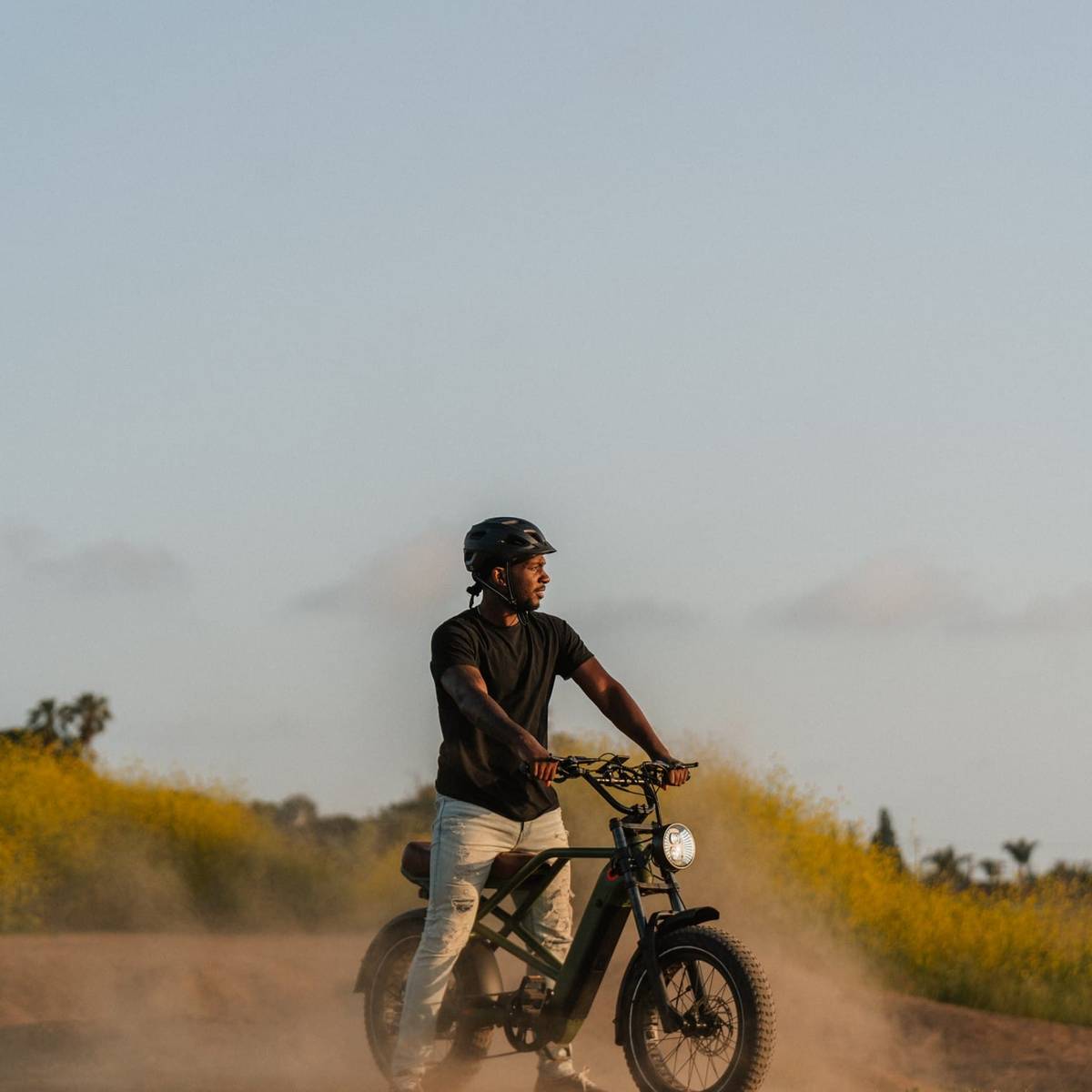 A person rides Valen Rev+ Fat Tire Electric Bike through a dusty landscape, with wildflowers blooming in the background under a clear sky.