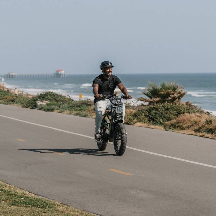 A person rides an olive green Valen Rev+ Fat Tire Electric Bike e along a coastal path with ocean waves and a pier visible in the background under a clear blue sky.