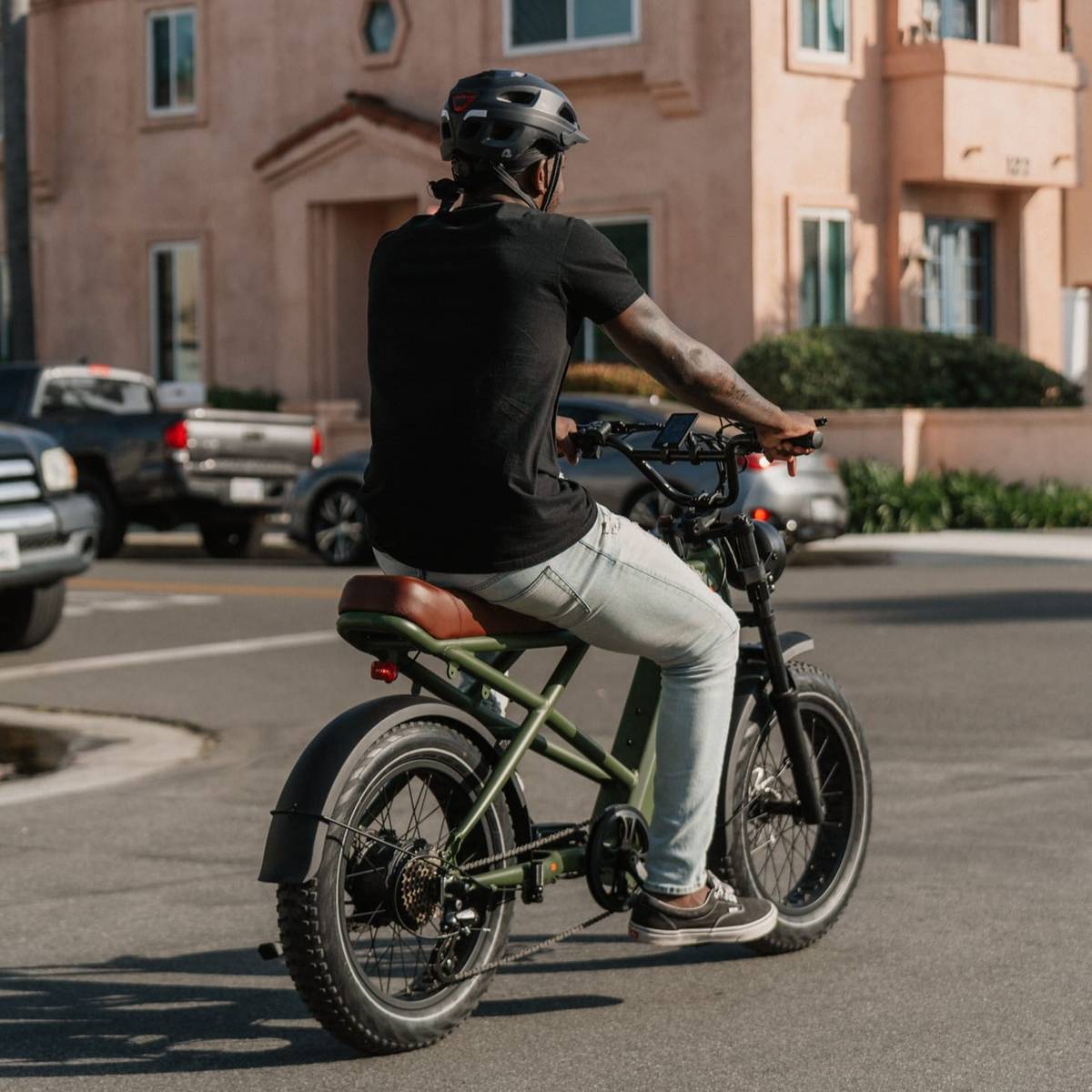 A person riding a matte olive green Valen Rev+ Fat Tire Electric Bike in a casual outfit and helmet, surrounded by cars and a pastel-colored building in the background.