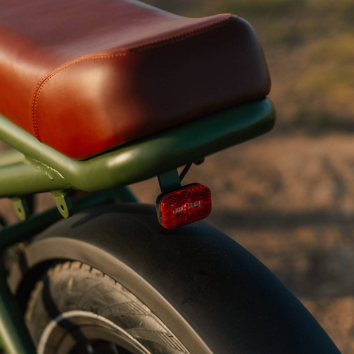 Close-up of the bike seat on the green Valen Rev+ Fat Tire Electric Bike with brown leather upholstery and a red reflector above the rear tire, set against a natural background.