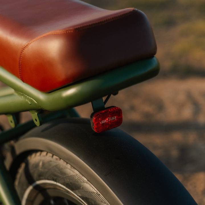 Close-up of the bike seat on the green Valen Rev+ Fat Tire Electric Bike with brown leather upholstery and a red reflector above the rear tire, set against a natural background.