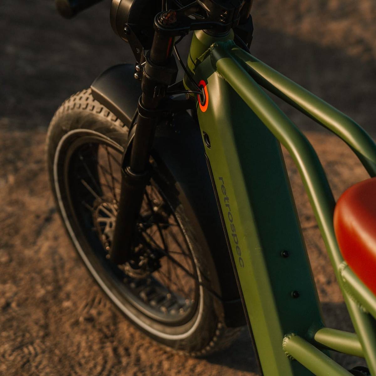 A close-up of a green Valen Rev+ Fat Tire Electric Bike frame with a brown seat and large black tires, resting on sandy ground in warm light.