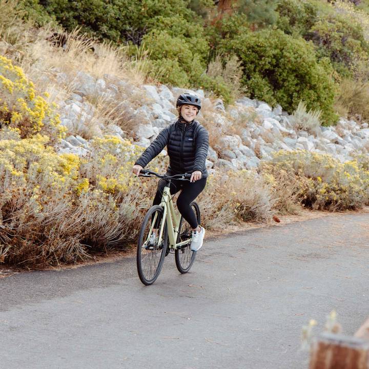 A person rides a light green bicycle on a pathway surrounded by yellow flowers and rocky terrain.