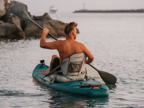 A man paddles while sitting on an inflatable kayak seat attached to the Weekender Tour iSUP on calm waters with rocks in the background.