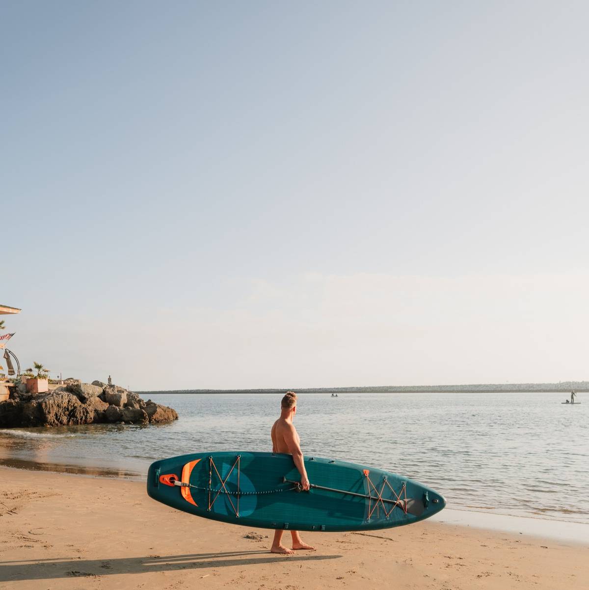 A person walks barefoot on a sandy beach, carrying a blue Weekender Tour Inflatable Stand Up Paddle Board, with calm waters and a jetty in the background.